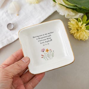 May include: A square, white ceramic trinket dish with a gold rim. The dish features a floral design and the text: "Having you as my mother is the greatest gift. I love you Mom Love, Sophia 10.8.25."