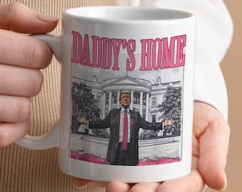 May include: White ceramic mug featuring a black and white illustration of a man in a suit standing in front of the White House. The text "DADDY'S HOME" is printed in pink letters on the mug.