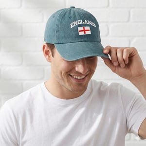 May include: A teal baseball cap with the word "ENGLAND" embroidered in white above a white and red English flag. The cap is made of a soft material and has a curved brim.