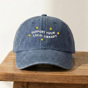 May include: A denim blue baseball cap with the text "SUPPORT YOUR LOCAL LIBRARY" in white, accented with yellow stars. The cap is sitting on a wooden surface.
