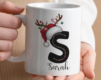 Peut inclure: Une tasse en céramique blanche avec une lettre noire "S" décorée de lumières de Noël colorées. Un chapeau de Père Noël rouge avec des bois de renne est posé sur la lettre. Le nom "Sarah" est écrit sous la lettre.