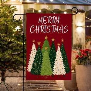 May include: A red garden flag with the words "MERRY CHRISTMAS" in white script. The flag features five Christmas trees in shades of green and white, adorned with gold stars and snowflakes. A festive outdoor decoration.