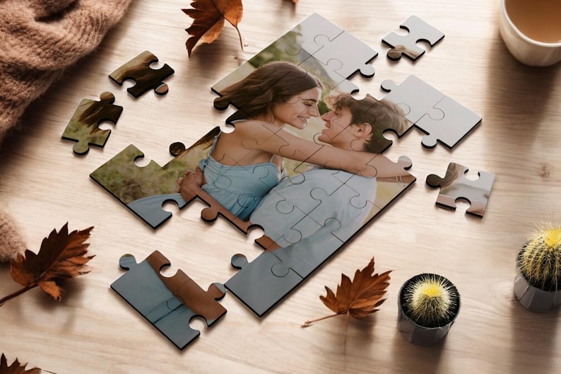 May include: A completed jigsaw puzzle with a photo of a couple embracing. The puzzle pieces are scattered around the image, with some pieces missing. The background is a wooden surface with fall leaves and a potted cactus.