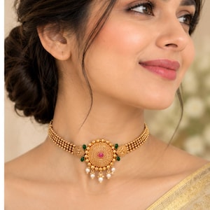 May include: A gold choker necklace featuring a central circular medallion with a small pink stone, accented by green teardrop-shaped stones on the sides and three dangling white pearls at the bottom, attached to a beaded gold chain.
