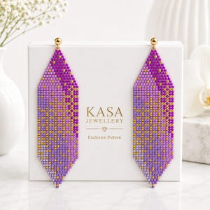 May include: A pair of long, diamond-shaped beaded earrings featuring a gradient of purple, lavender, and gold beads. The earrings are displayed on a white box labeled KASA JEWELLERY Exclusive Pattern with a small diamond icon centered below.