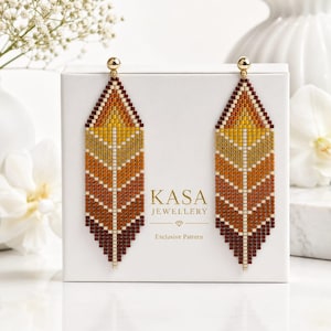 May include: A pair of long, leaf-shaped beaded earrings featuring a gradient of yellow, orange, and dark red beads. The earrings are displayed on a white box labeled KASA JEWELLERY Exclusive Pattern with a small diamond icon centered below.
