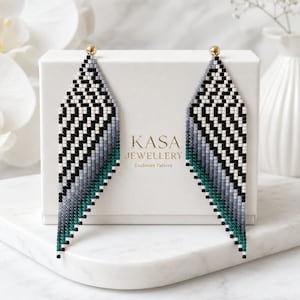 May include: A pair of long, triangular beaded earrings featuring a geometric chevron pattern in black, white, grey, and teal. The earrings are displayed against a white box labelled KASA JEWELLERY Exclusive Pattern with gold stud tops.