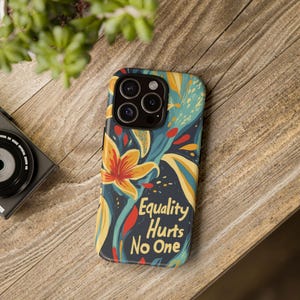 May include: A phone case with a floral design in yellow, orange, and teal on a dark blue background. The case has the text "Equality Hurts No One" in red and yellow. The phone case is on a wooden surface.