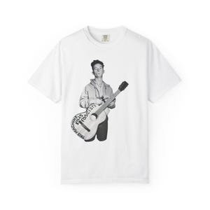 May include: White t-shirt featuring a black and white image of a person holding a guitar. The guitar has the text "THIS MACHINE KILLS FASCISTS" printed on it. The shirt has a crew neck and short sleeves.