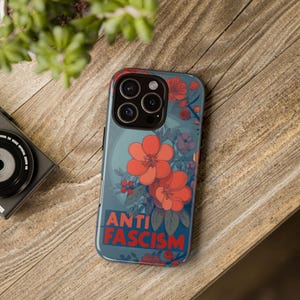 May include: A phone case with a floral design and the words "ANTI FASCISM" in red. The case features large orange flowers, smaller purple berries, and green leaves against a teal background. The case is on a wooden surface.