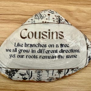 Puede incluir: Concha decorativa con la palabra "Cousins" en letras marrones. La concha tiene un fondo blanco con diseños florales negros en los bordes. También está presente el texto "Como ramas en un árbol, todos crecemos en diferentes direcciones, pero nuestras raíces permanecen iguales".