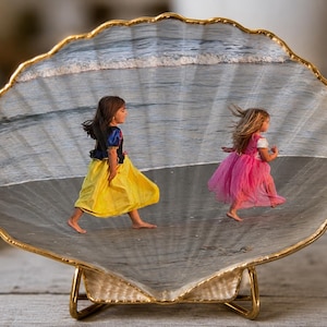 Op de afbeelding: Een decoratieve sint-jakobsschelp met een afgedrukte foto van twee kinderen die op een strand rennen. Eén kind draagt een geel met blauwe jurk, de ander een roze jurk. De schelp heeft een goudkleurige rand en staat op een gouden standaard.