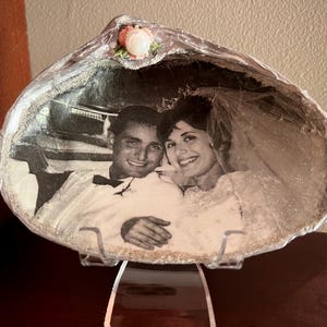 May include: A large, natural seashell serving as a frame for a black and white wedding photograph. The shell features a silver-painted rim and a small, decorative pink and white floral accent at the top centre, displayed on a clear plastic stand.