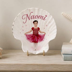 May include: A white scallop shell featuring a printed photograph of a child in a red ballet outfit with a tutu. The name Naomi is written in pink cursive script above the child, surrounded by small pink star icons on the shell surface.