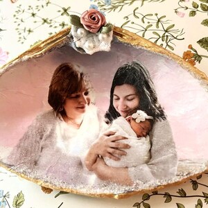 Può includere: Una conchiglia decorativa con bordo dorato, con una foto di famiglia. La foto mostra due donne e un bambino. Il guscio è adornato con una piccola composizione floreale in cima, aggiungendo un tocco delicato al ricordo.