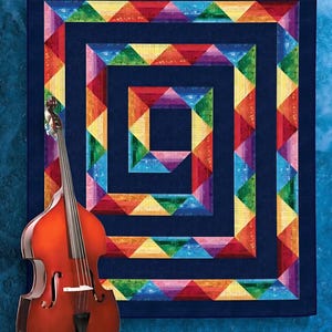 May include: A colorful quilt with a rainbow geometric design against a navy blue background. A wooden double bass leans against the wall. The quilt features a square-in-square pattern with vibrant triangles in red, orange, yellow, green, and purple.
