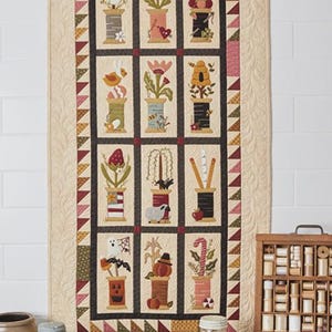 Puede incluir: Un edredón decorativo con carretes ilustrados con diseños estacionales y caprichosos. El edredón mide 61 cm por 124 cm y se exhibe contra una pared blanca. El edredón es de Buttermilk Basin Design Co.