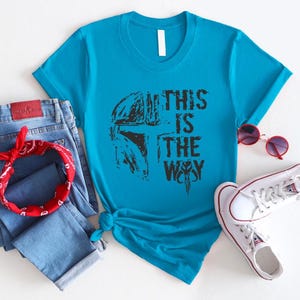 May include: A turquoise t-shirt with the text "THIS IS THE WAY" in black, paired with blue jeans, a red bandana, and white sneakers. The shirt features a graphic design.