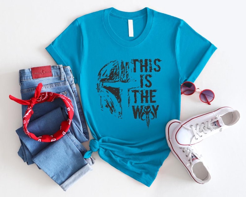 May include: A turquoise t-shirt with the text "THIS IS THE WAY" in black, paired with blue jeans, a red bandana, and white sneakers. The shirt features a graphic design.