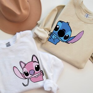 K&ouml;nnte beinhalten: Wei&szlig;e und beige Sweatshirts mit Rundhalsausschnitt und Cartoon-Charakter-Grafiken. Das wei&szlig;e Sweatshirt zeigt eine rosa Figur, w&auml;hrend das beige Sweatshirt eine blaue Figur zeigt. Im Hintergrund ist ein brauner Hut zu sehen.