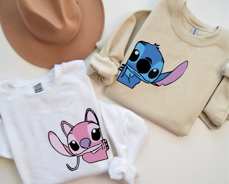 K&ouml;nnte beinhalten: Wei&szlig;e und beige Sweatshirts mit Rundhalsausschnitt und Cartoon-Charakter-Grafiken. Das wei&szlig;e Sweatshirt zeigt eine rosa Figur, w&auml;hrend das beige Sweatshirt eine blaue Figur zeigt. Im Hintergrund ist ein brauner Hut zu sehen.
