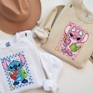 Puede incluir: Dos sudaderas, una blanca y otra beige, cada una con un personaje de dibujos animados. La sudadera blanca tiene un personaje azul con un libro rojo y una mochila verde. La sudadera beige tiene un personaje rosa con un libro verde y un l&aacute;piz.