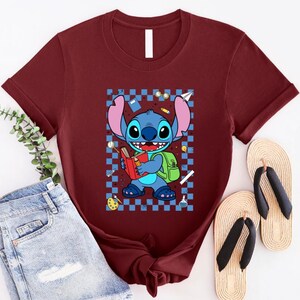 Puede incluir: Camiseta granate con una ilustraci&oacute;n de dibujos animados de Stitch de Lilo & Stitch. Stitch sostiene un libro y lleva una mochila, sobre un fondo a cuadros azules y blancos. La imagen incluye un par de chanclas.