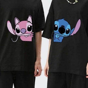 K&ouml;nnte beinhalten: Schwarze T-Shirts mit den Zeichentrickfiguren Angel und Stitch. Angel ist rosa und Stitch ist blau. Beide Charaktere sind teilweise auf den Shirts zu sehen. Die Shirts haben kurze &Auml;rmel und eine lockere Passform.