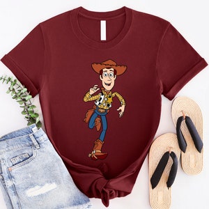 May include: A maroon t-shirt with a cartoon image of Woody from Toy Story. Woody is wearing his signature cowboy hat, vest, and jeans. He is holding a lasso in his right hand and has a friendly expression on his face.