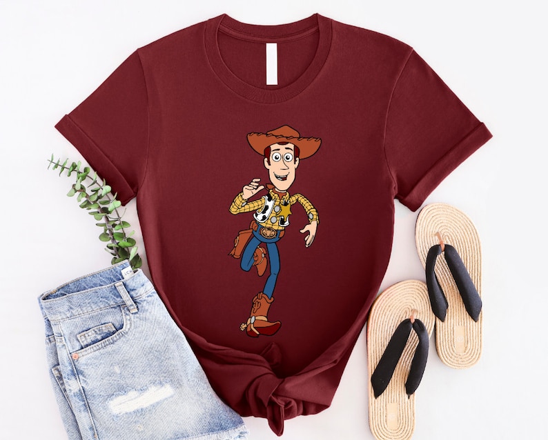 May include: A maroon t-shirt with a cartoon image of Woody from Toy Story. Woody is wearing his signature cowboy hat, vest, and jeans. He is holding a lasso in his right hand and has a friendly expression on his face.