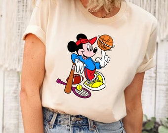 May include: A white t-shirt with a cartoon image of Mickey Mouse playing sports. Mickey is wearing a red hat, blue shirt, and red pants. He is holding a basketball and there are other sports equipment around him, including a baseball bat, tennis racket, baseball, and tennis ball.