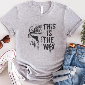 May include: A gray t-shirt with the phrase "THIS IS THE WAY" printed in black, along with a helmet graphic. The shirt is paired with blue jeans, white sneakers, and sunglasses.