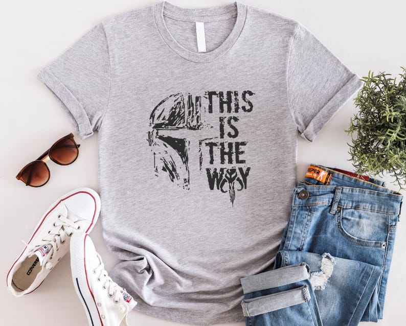 May include: A gray t-shirt with the phrase "THIS IS THE WAY" printed in black, along with a helmet graphic. The shirt is paired with blue jeans, white sneakers, and sunglasses.