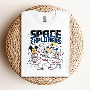 以下が含まれることがあります： 宇宙飛行士の衣装を着たミッキーマウス、グーフィー、ドナルドダックが描かれた白いTシャツ。「SPACE EXPLORERS」の文字が黒と青でプリントされています。Tシャツは、織られた円形の表面に折りたたまれています。