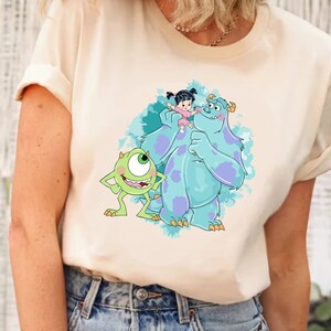 May include: A white t-shirt with a cartoon image of Sully, Mike, and Boo from the movie Monsters Inc. Sully is blue and has purple spots. Mike is green with one large eye. Boo is a small girl with black hair and a pink dress.
