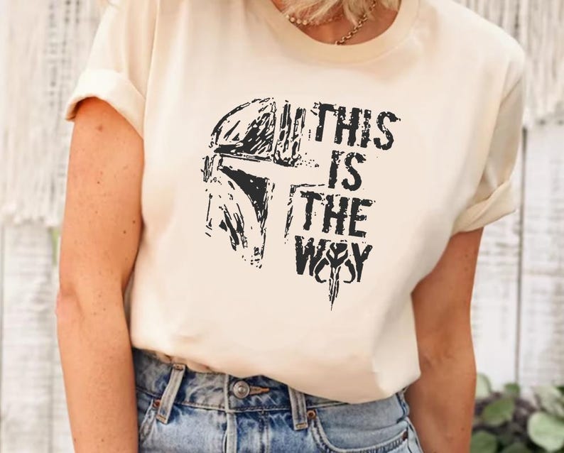 May include: Cream-colored t-shirt featuring a black graphic of a helmet and the text "THIS IS THE WAY." The design is a distressed style. The shirt has short sleeves and a crew neck.