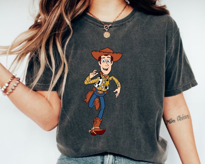May include: A dark gray t-shirt with a cartoon image of Woody from Toy Story. Woody is wearing his signature cowboy hat, vest, and jeans. He is holding a toy sheriff's badge in his right hand and a toy gun in his left hand. The t-shirt has a distressed look.