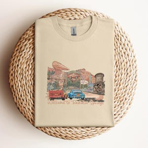 May include: A beige t-shirt featuring a colorful illustration of a desert town with vintage cars and buildings. The text "Welcome to Radiator Springs" is printed below the image. The shirt is folded on a woven, circular surface.