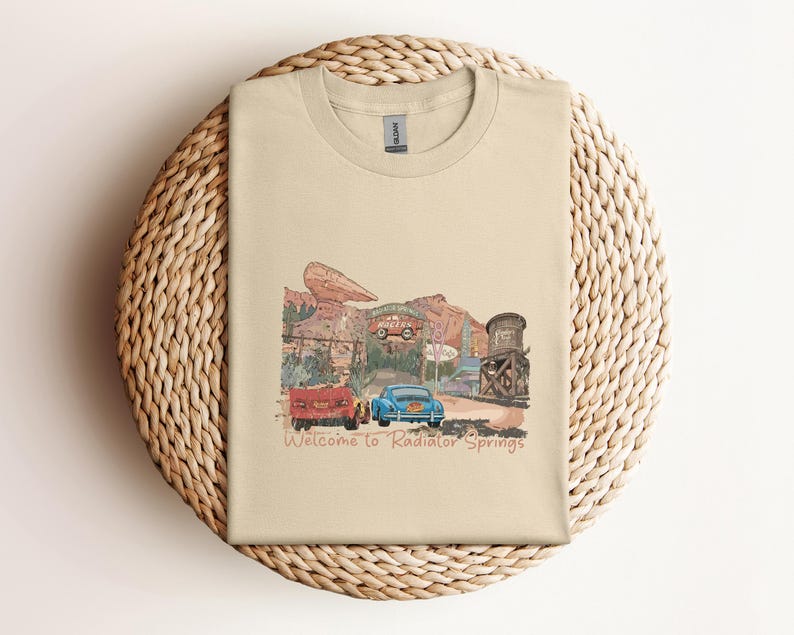May include: A beige t-shirt featuring a colorful illustration of a desert town with vintage cars and buildings. The text "Welcome to Radiator Springs" is printed below the image. The shirt is folded on a woven, circular surface.
