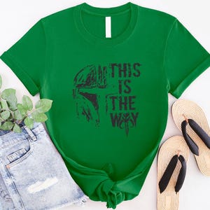 May include: A bright green t-shirt featuring a black graphic of a helmet and the text "THIS IS THE WAY." The shirt is styled with denim shorts and sandals.