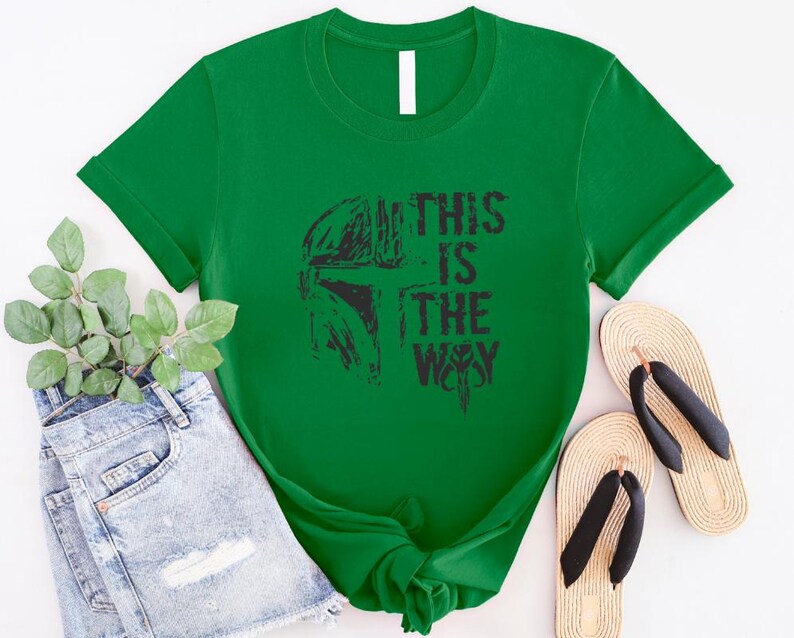 May include: A bright green t-shirt featuring a black graphic of a helmet and the text "THIS IS THE WAY." The shirt is styled with denim shorts and sandals.
