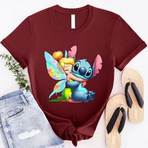 K&ouml;nnte beinhalten: Ein maroonfarbenes T-Shirt mit einem Cartoon-Bild von Stitch aus Lilo & Stitch, der Tinkerbell aus Peter Pan umarmt. Stitch ist blau und Tinkerbell tr&auml;gt ein gr&uuml;nes Kleid und Feenfl&uuml;gel mit blauen, rosa und gelben Akzenten.