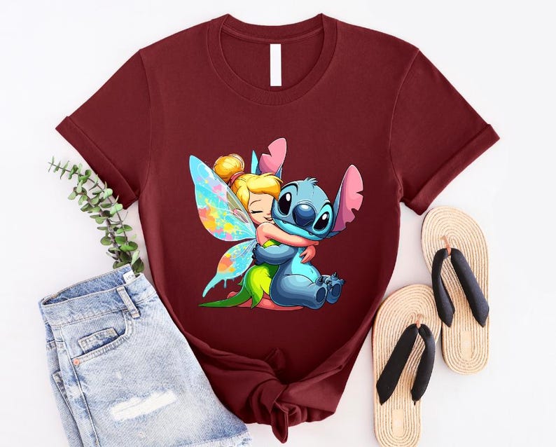 K&ouml;nnte beinhalten: Ein maroonfarbenes T-Shirt mit einem Cartoon-Bild von Stitch aus Lilo & Stitch, der Tinkerbell aus Peter Pan umarmt. Stitch ist blau und Tinkerbell tr&auml;gt ein gr&uuml;nes Kleid und Feenfl&uuml;gel mit blauen, rosa und gelben Akzenten.