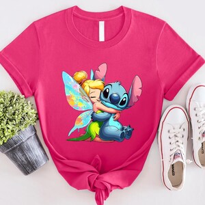 K&ouml;nnte beinhalten: Pinkes T-Shirt mit einem Cartoon-Bild von Stitch aus Lilo & Stitch, der Tinkerbell aus Peter Pan umarmt. Das Bild ist im farbenfrohen Cartoon-Stil gehalten.
