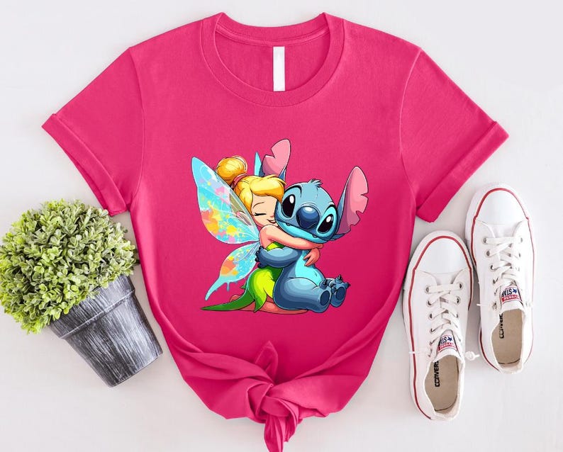 K&ouml;nnte beinhalten: Pinkes T-Shirt mit einem Cartoon-Bild von Stitch aus Lilo & Stitch, der Tinkerbell aus Peter Pan umarmt. Das Bild ist im farbenfrohen Cartoon-Stil gehalten.