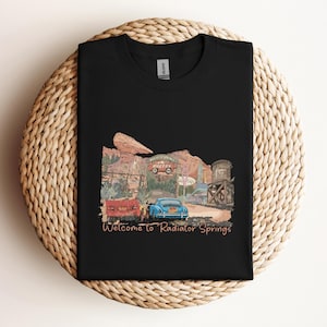 May include: Black t-shirt featuring a colorful graphic of a desert town scene with vintage cars and the text "Welcome to Radiator Springs." The design includes a red truck, a blue car, and a water tower. The shirt is displayed on a woven, circular surface.