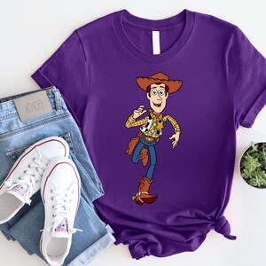 May include: Purple t-shirt with a cartoon image of Woody from Toy Story. Woody is wearing his signature cowboy hat, vest, and jeans. He is holding his lasso and smiling.