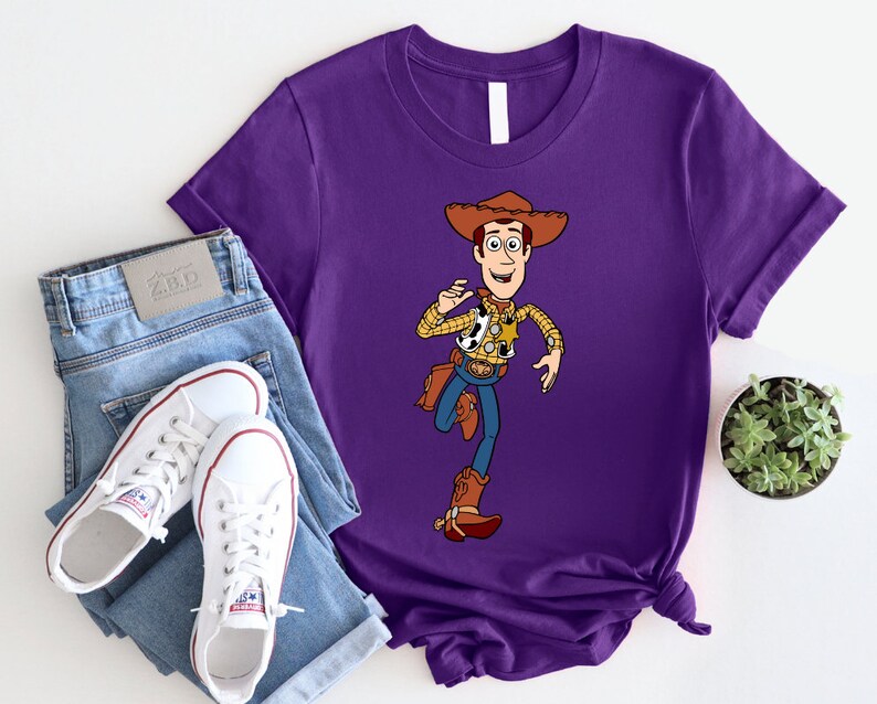 May include: Purple t-shirt with a cartoon image of Woody from Toy Story. Woody is wearing his signature cowboy hat, vest, and jeans. He is holding his lasso and smiling.