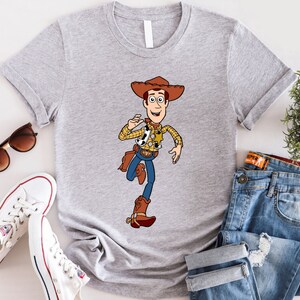 May include: A gray t-shirt with a cartoon image of Woody from Toy Story. Woody is wearing his signature cowboy hat, vest, and boots. He is holding a sheriff's badge and has a friendly smile on his face.
