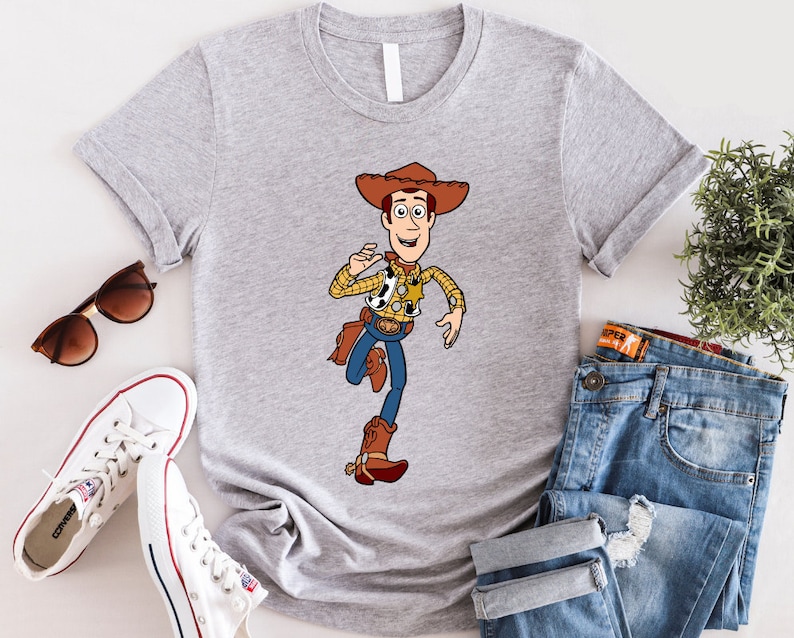 May include: A gray t-shirt with a cartoon image of Woody from Toy Story. Woody is wearing his signature cowboy hat, vest, and boots. He is holding a sheriff's badge and has a friendly smile on his face.